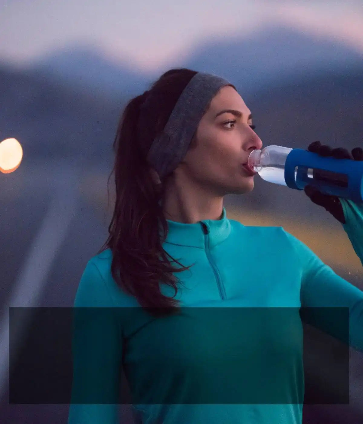 Someone in a turquoise athletic top drinking from a water bottle at dusk.