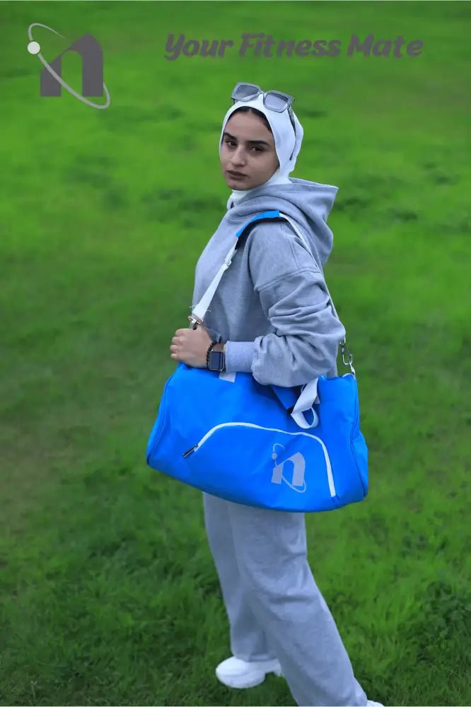 Person wearing a light gray tracksuit and white hijab carrying a bright blue sports bag.